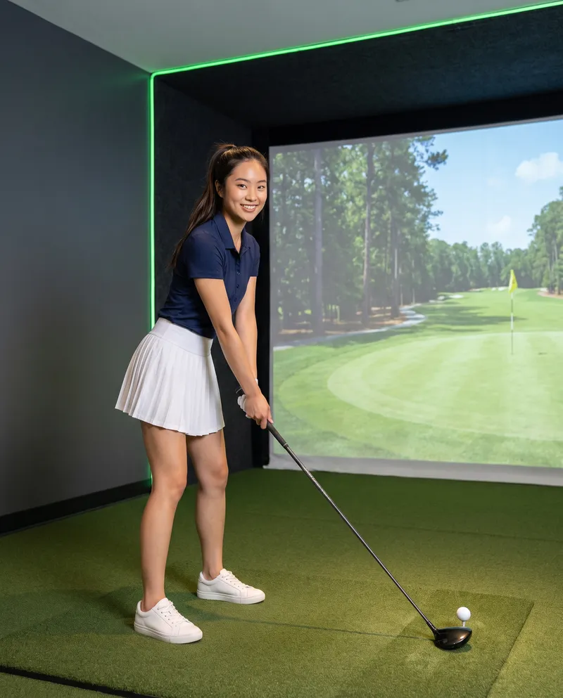 Generated picture of the same character at address with a driver on the hitting mat of a simulator bay, smiling at the camera, a fairway and a green with a flag on the screen behind her.